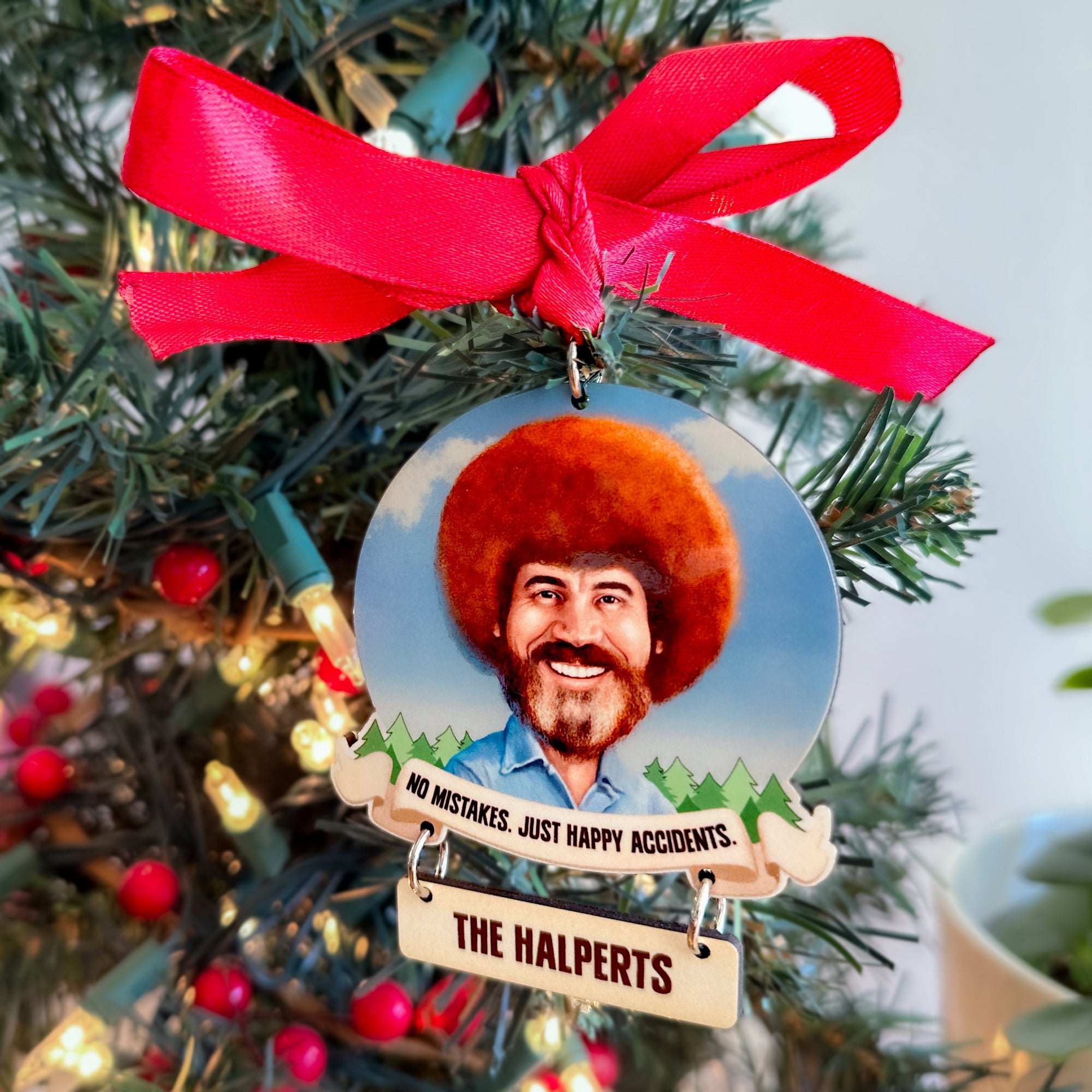 Christmas ornament with a portrait of Bob Ross and a red ribbon, hanging on a decorated Christmas tree.